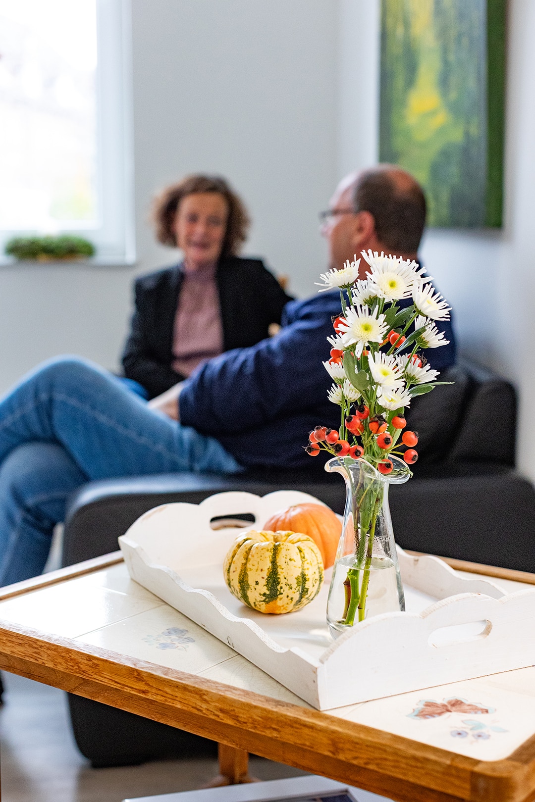 Blumen und Kürbisse auf Tisch, dahinter entspanntes Gespräch zwischen zwei Personen in gemütlichem Wohnzimmerumfeld.